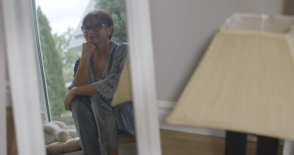 Reflection in Mirror of Stylish Confident Beautiful Senior Woman in Eyeglasses Sitting on Windowsill