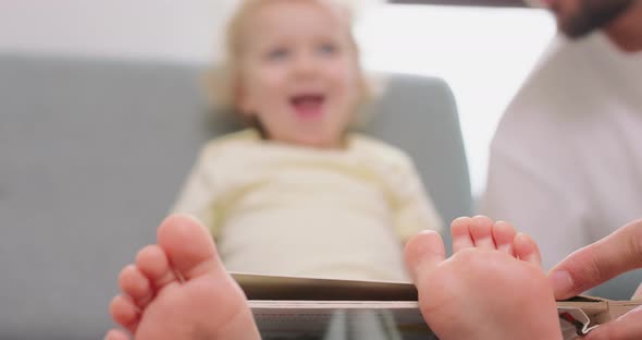 Closeup of a Little Girl's Feet on a Gray Sofa Who is Looking at Pictures in a Book with Her Dad Her