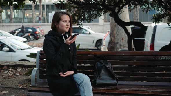 Portrait of a Smiling Hipster Girl Typing Message on Mobile Phone While Sitting on Bench at Park