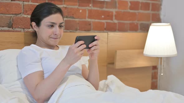 Young Indian Woman Playing Video Game on Smartphone in Bed