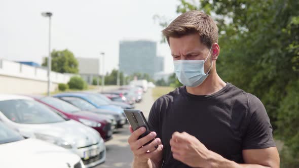 A Young Handsome Man in a Face Mask Works on a Smartphone in a Parking Lot in an Urban Area