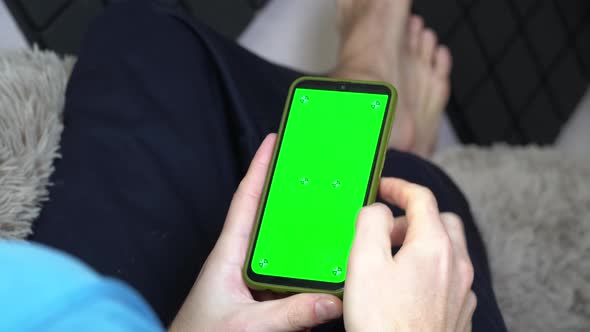 A Man on the Couch With a Phone in His Hands With a Green Screen Back View