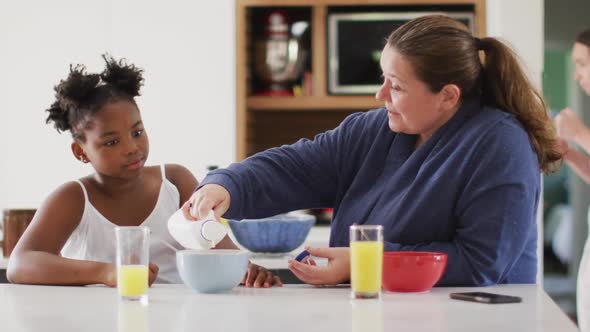 Happy caucasian lesbian couple and their african american daughter eating breakfast in kitchen