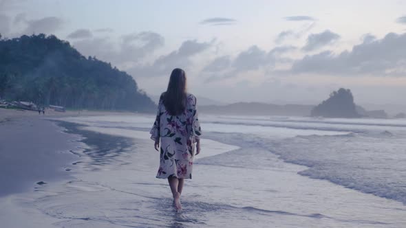 Young brunette woman walking away from camera on long Asian beach at cloudy dusk in slow motion