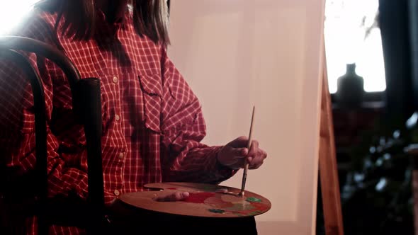 A Young Pretty Woman Artist Mixing Warm Colors on the Palette Sitting in Front of an Easel