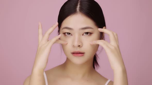 Close up face of beautiful young Asian woman with clear skin lifting eye on pink background.