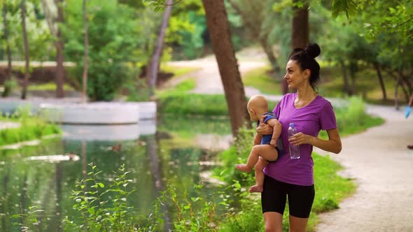 Fit Woman Carrying Baby and Drinking Water After Training in Park