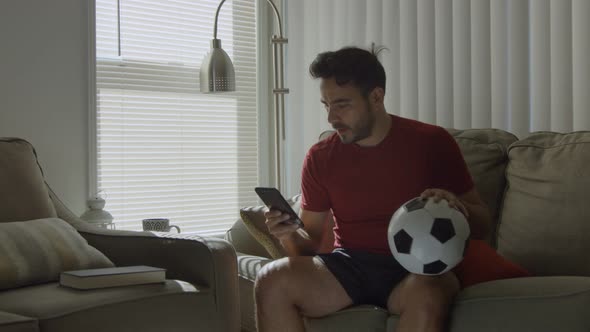 Man Watching Soccer Game on Cellphone While Holding Soccer Ball at Home