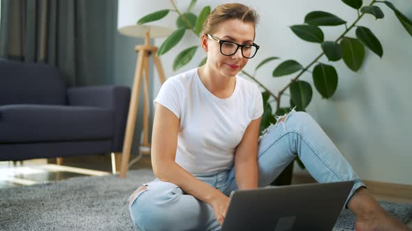 Woman with Glasses is Sitting on the Floor and Working on a Laptop