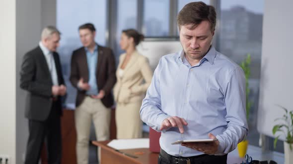 Angry Male Employee Messaging Online on Tablet in Office with Blurred Group of Colleagues Talking at