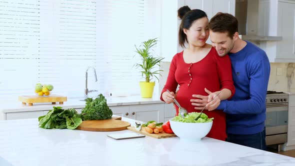 Man embracing pregnant woman while preparing salad at kitchen counter 