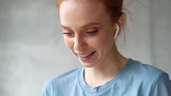 Close-up of Face of Cheerful Redhead Young Woman Wearing Wireless Earphones Watching Video on Phone.