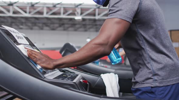 Fit african american man wearing face mask and headphones cleaning treadmill machine with disinfecta