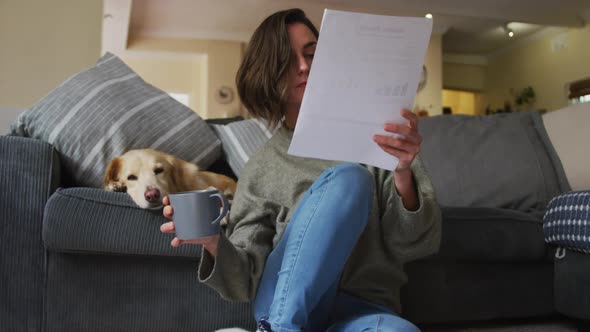 Caucasian woman reading documents, working from home with her pet dog on sofa next to her