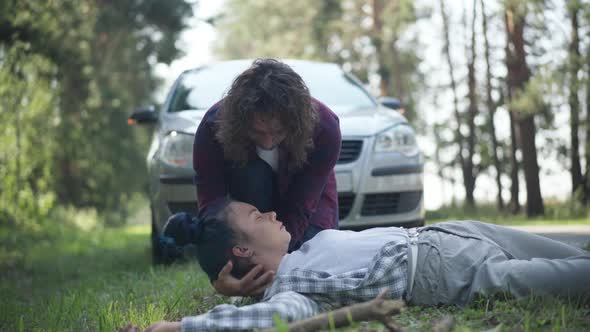 Portrait of Young Man Checking Pulse of Knocked Down Woman with Car at Background on Suburban