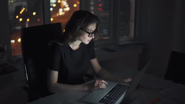 Portrait of a Young Girl Working Late in the Office. Business Woman in Glasses Works on a Laptop on