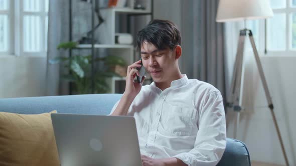 Smiling Asian Man Answering The Call On Smartphone While Lying On Sofa And Using Laptop Computer