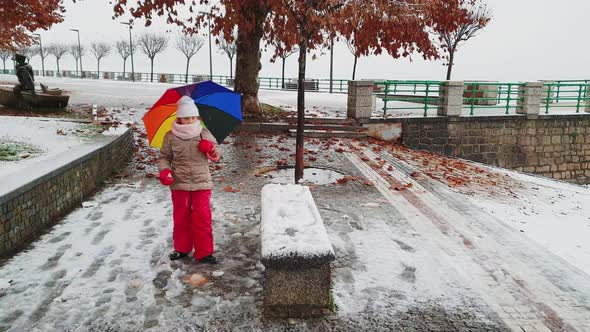 Little child girl with umbrella of many colors plays kicking snowball. Slow-motion