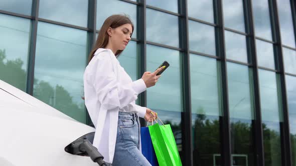 Young Girl After Buying Products with a Shopping Bag is Standing Near the Charging Electric Car