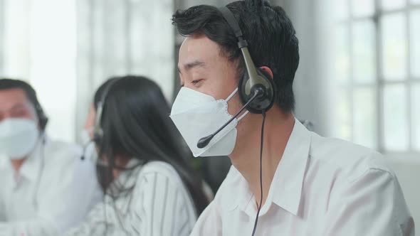 Man Of Three Asian Call Centre Agents In Mask Speaking To Customer While His Colleagues Are Talking