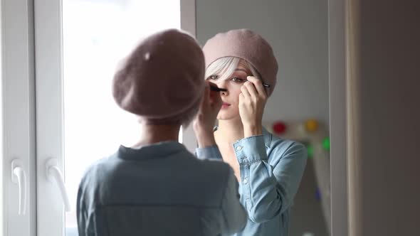 Young blonde woman applying makeup at home near a mirror.