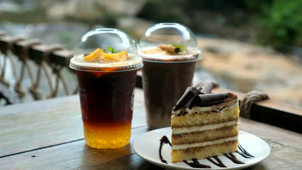 Two Cups with Cold Drinks and Plate with Piece of Chocolate Cake Standing on Wooden Table in Outdoor