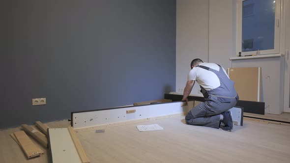A Builder in a Work Overalls is Assembling a Bed in a New Apartment