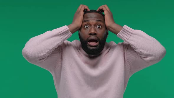 Shocked African American Man with Open Mouth Holding His Head Over Green Background in the Studio