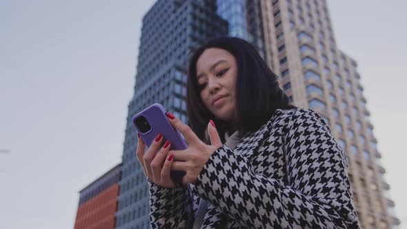 Young Adult Asian Woman Browsing on Her Smartphone Under a Highrise Building