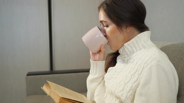 Pretty Girl Reading a Book and Drinking Coffee on the Sofa at Home