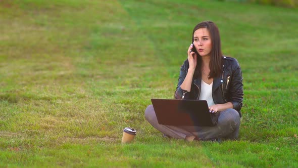 A Girl in the Park on the Lawn Working on a Laptop, Phone Rings, Answers an Incoming Call