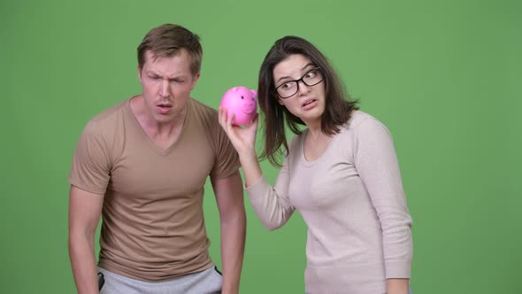 Young Couple Shaking Piggy Bank Together Against Green Background