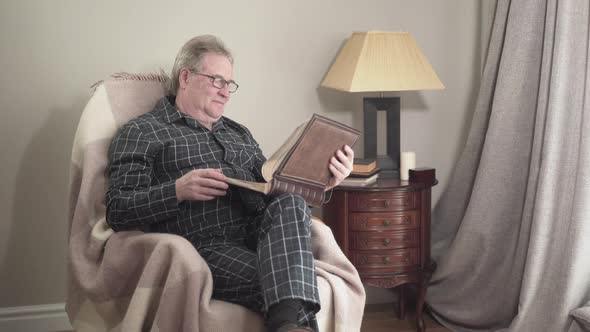 Old Caucasian Man in Pajamas Sitting in Rocking Chair and Looking at Photos in Photo Album. Positive