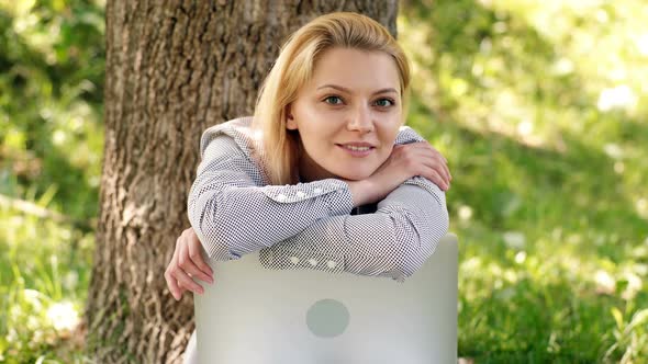 Young Woman Sits on a Grass in a Park Hold Laptop and Smiles. Dreamy Female Student.