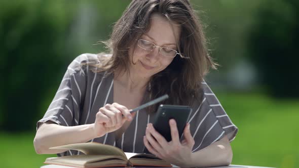 Pretty Adult Woman is Reading Book in Foreign Language and Using Smartphone for Translating