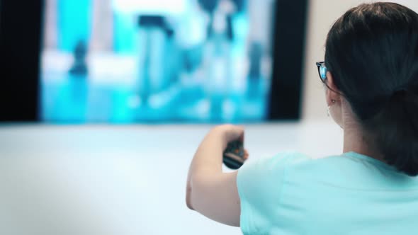 Cinematic Brunette Teen Girl with Glasses Sits on the Sofa in Front of the TV and Switches Channels