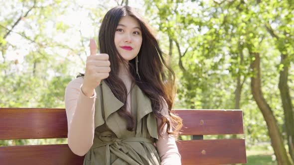 Young Asian Woman Shows Thumb Up To Camera Nods Smile As She Sits Bench