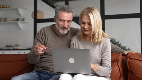 Cheerful Middleaged Couple Watching at Laptop Screen and Checking Credit Card Number
