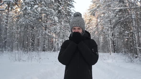 Portrait young man in knitted hat and mittens warms face. Frosty day in forest