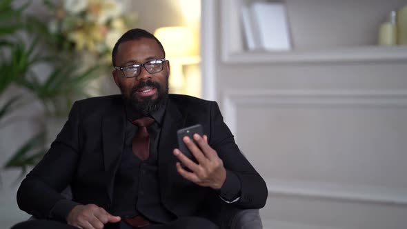 African-American with a Beard, Black Suit, Stylish Glasses. The Businessman Is Sitting on a Chair