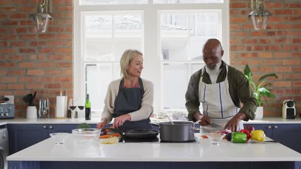 Mixed race senior couple wearing aprons cooking food together in the kitchen at home