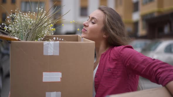 Portrait of Young Caucasian Brunette Woman with Heavy Boxes Outdoors on Parking Lot at Residential