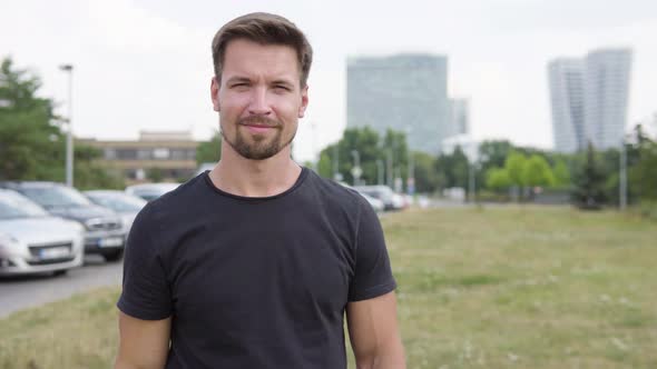 A Young Man Shows a Thumb Up To the Camera with a Smile By a Parking Lot - a City in the Background