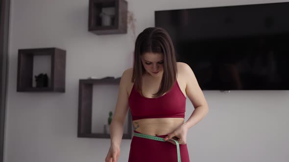 Fitness Woman in Sportswear Measures Waist Circumference with Tape in the Kitchen Feels Upset