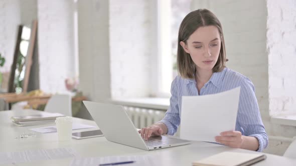 Focused Young Woman Reading Documents and Working on Laptop