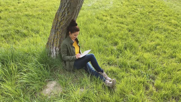 Young woman writing in notebook, sitting on meadow