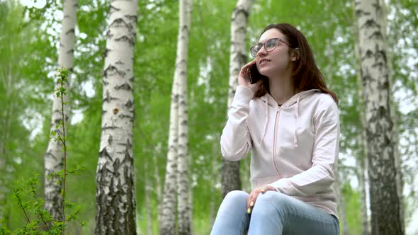 Young Woman in Nature with a Phone. A Girl Sits on a Stump in a Birch Forest and Talks on the Phone.