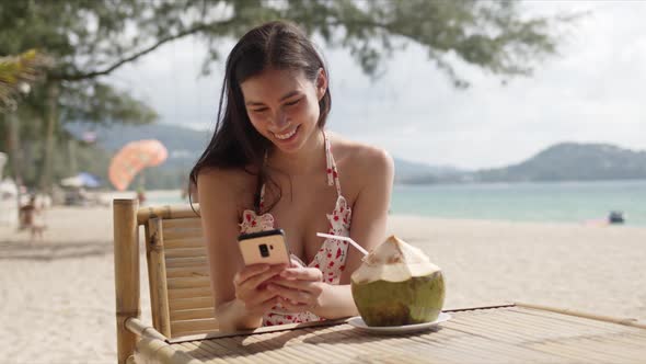 Young Dark Haired Female in Summer Dress Checking Message on Smartphone While Sitting in Beach Cafe