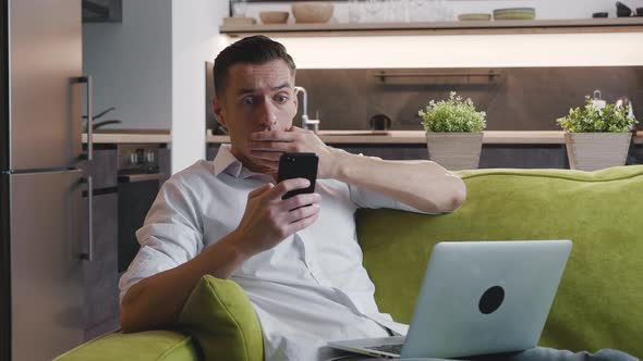 Shocked Man Reacting Bad Message on Smartphone Covers His Mouth in Fright Sitting on Sofa with