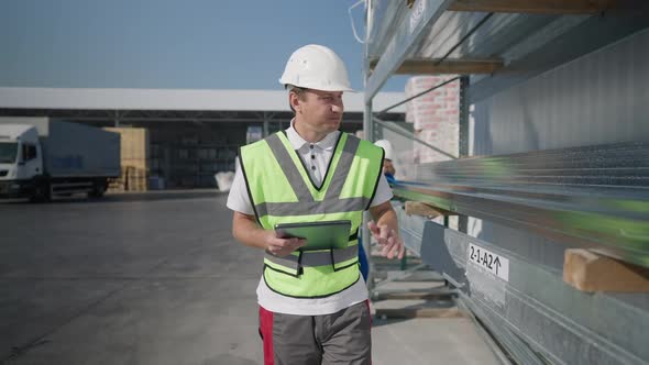 Confident Serious Caucasian Man in Hard Hat Walking Along Row of Delivered Metal Products at